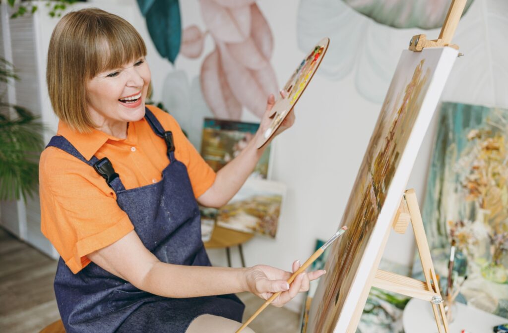 A senior woman smiling widely as she enjoys painting on a canvas.