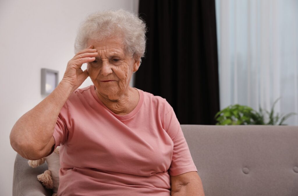 An elderly woman sitting on a couch with her hand held against her forehead as she struggles to remember something she needed to do.